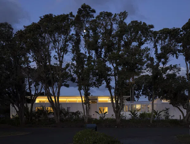 A low, single-storey modern building photographed at dusk, partially screened by mature trees and landscaped planting. The long façade has warm illuminated glazing or recessed lighting, pale walls, horizontal louvres, and a flat roofline, with a dark paved foreground and a small covered entry or carport element at the right.