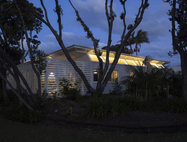 Twilight exterior view of a low, contemporary building partially screened by mature trees and tropical planting. The building has a shallow layered roofline with warm concealed lighting, pale horizontal louvred or slatted screening across the facade, and light-coloured walls. The scene is dark and atmospheric, with the illuminated roof and facade contrasting against the blue evening sky.