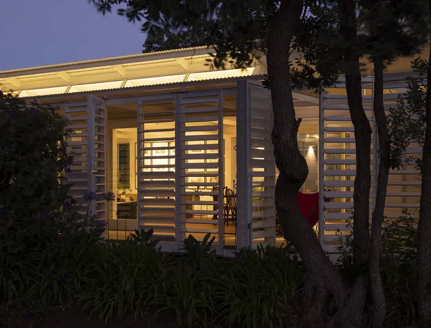 Twilight exterior view of a low-rise building screened by mature trees and dense planting. The facade features repeated horizontal timber-like slatted screens, glazed openings, and warm interior lighting. A shallow corrugated roof with a broad projecting canopy is visible, creating a layered, sheltered entry or outdoor transition zone.