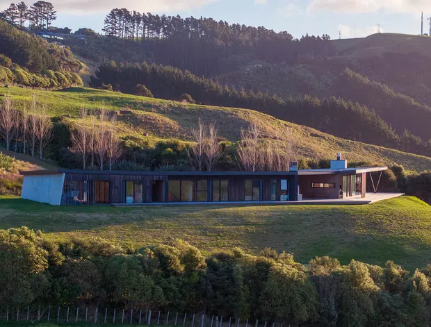 A low, elongated single-storey modern residence set into a broad rural hillside. The building has dark timber or metal-clad walls, extensive floor-to-ceiling glazing, a shallow roofline, and a projecting open-sided terrace or entry canopy. Rolling green pasture, hedgerows, and dense forested hills surround the home.