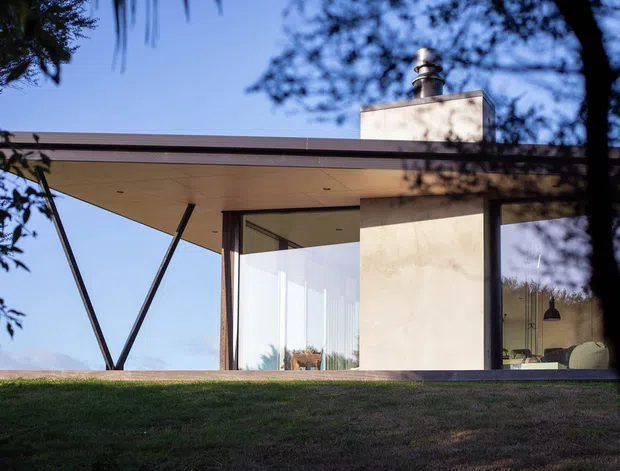 Exterior view of a contemporary low-profile house set behind a grassy foreground, with broad floor-to-ceiling glazing, slim dark framing, a deep flat roof overhang, and a prominent rendered chimney volume. Interior furnishings are faintly visible through the glass, while trees frame the view.