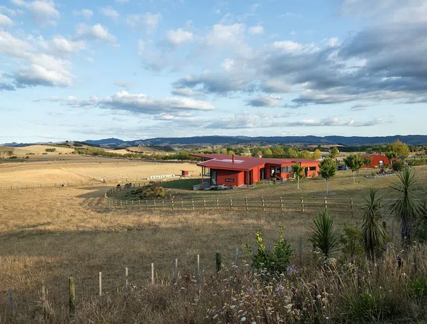 A low, modern red-clad rural building set within expansive dry agricultural fields, with rolling hills and a dramatic partly cloudy sky in the background. The property includes landscaped grounds, fencing, and several connected or adjacent structures.