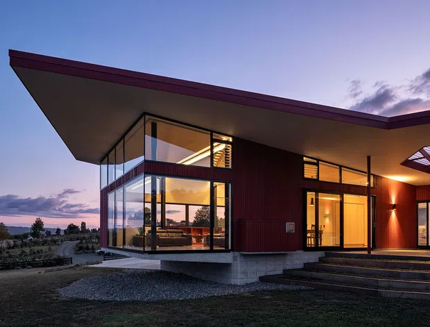 Contemporary single-storey house photographed at dusk, with a dramatic low-pitched overhanging roof, extensive floor-to-ceiling glazing, dark red vertical cladding, concrete base, and warm interior lighting. Broad steps lead to the entry, while the glazed corner reveals a living area and a sculptural staircase. The property sits in an open landscaped setting with distant water or horizon views.
