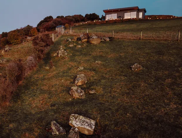 A small contemporary single-storey house sits on a gently sloping rural hillside. The building has a simple rectangular form, broad glazed openings, pale walls, and a low-pitched dark roof with a projecting roof edge. Fenced grassland, scattered rocks, and mature vegetation surround the house, photographed in warm low-angle light.