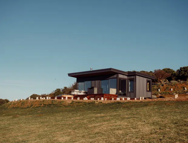 A compact modern single-storey dwelling set on an open grassy site. The house has dark vertical cladding, extensive floor-to-ceiling glazing, a low asymmetrical flat or gently sloped roof, and a raised masonry base with a small outdoor seating area. Rocky landscaped terrain and scrub surround the building.