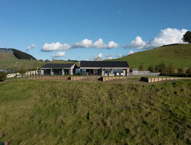 Wide exterior view of a low, modern single-storey building set into a gently sloping rural landscape. The building has a dark pitched roof, extensive glazing, pale walls, covered outdoor areas, and a long timber-toned deck or paved terrace. Rolling grassy hills and scattered rural development surround the property under a bright blue sky.