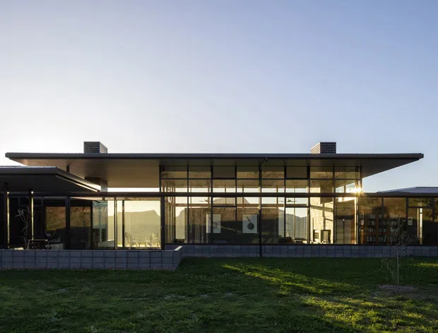 Single-storey modern residence viewed from the exterior at dusk, with a broad low-pitched roof and deep overhangs extending across glazed living spaces. Floor-to-ceiling glazing forms a transparent central facade, with dark slim frames, masonry base walls, and smaller projecting wings on either side. The building sits in an open grassed setting with warm sunlight reflecting through the glazing.