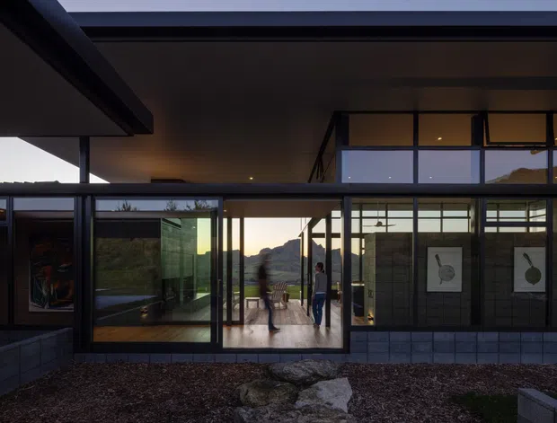A low-slung modern residential building viewed from a landscaped courtyard, with extensive floor-to-ceiling glazing, dark slender window frames, covered outdoor circulation, and a partially visible interior. The structure opens toward a distant hilly landscape, with warm interior lighting contrasting against the dusky exterior.