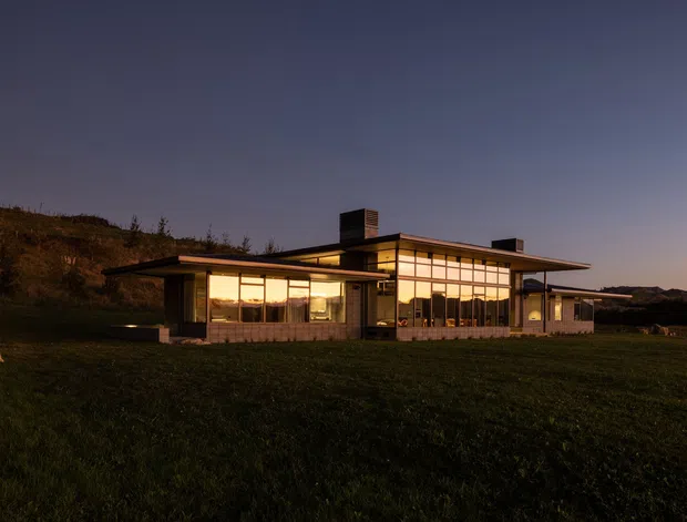 A low, single-storey modern house photographed at dusk, set within an open grassy landscape. The building has broad horizontal roof planes with deep overhangs, extensive floor-to-ceiling glazing, slender dark structural framing, warm interior lighting, and prominent chimney elements. The surrounding terrain appears gently sloped and rural.