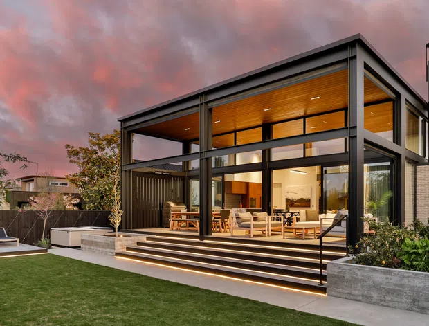 Contemporary two-storey residential house viewed from the landscaped rear garden at dusk. The home has a strong rectilinear form, dark metal-framed floor-to-ceiling glazing, a projecting upper-level volume, warm timber soffits and ceilings, exposed outdoor steps, and a covered entertaining terrace. A lawn, raised concrete planter, boundary fencing, and outdoor seating occupy the foreground.