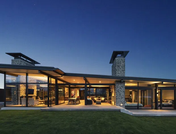 Wide dusk exterior view of a low-slung, single-storey contemporary residence with extensive floor-to-ceiling glazing, broad projecting roof planes, dark slender structural framing, and prominent stone-clad chimneys. Warm interior lighting reveals living and dining spaces through the glass, while a manicured lawn and paved terrace occupy the foreground.