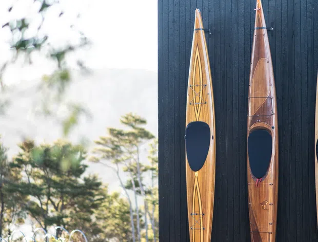 Exterior detail showing three vertically mounted wooden kayaks displayed against a dark vertical timber-clad wall, with blurred trees and bright outdoor light visible to the left.