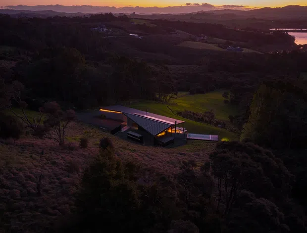 Aerial dusk view of a contemporary low-profile home nestled into a heavily vegetated, rolling rural or bush landscape. The building has angular wings, dark exterior finishes, broad roof planes, and warm interior lighting, with distant hills and a reflective body of water visible on the horizon.
