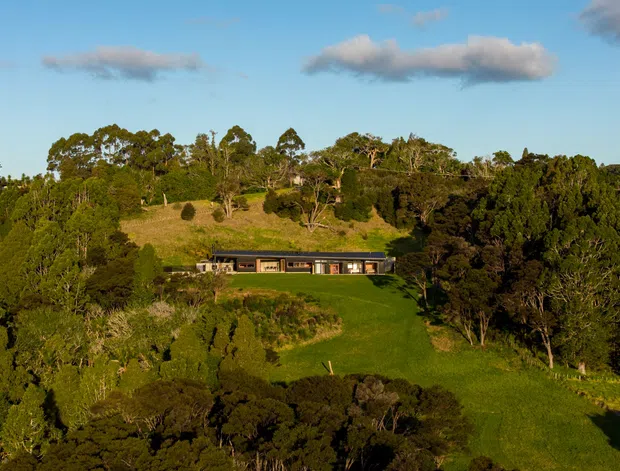 Wide exterior landscape view of a low, elongated modern home set into a large grassy clearing on a densely wooded, gently elevated site. The house has a dark, low-pitched or flat roof, extensive horizontal glazing, and a restrained linear form. The surrounding mature trees and open lawn dominate the scene under a clear blue sky.