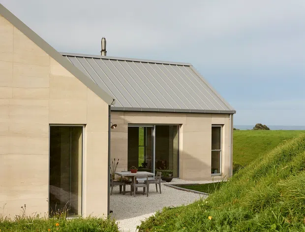 Exterior view of a minimalist coastal house set into a grassy landscape. The building has pale rendered or concrete walls, a simple pitched standing-seam metal roof, large glazed openings, and a gravel courtyard with an outdoor dining table. A narrow pale path leads through the surrounding grass toward the house, with the sea visible in the distance.