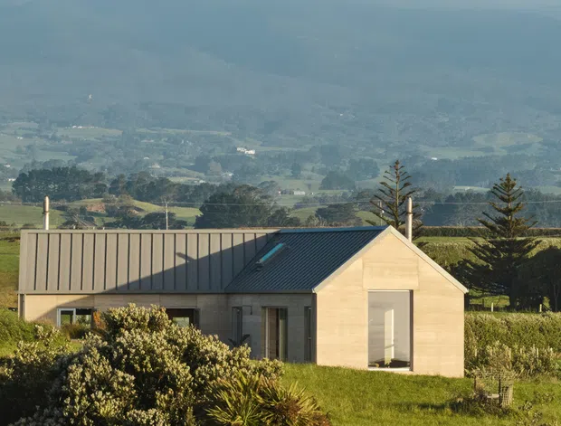 A compact modern rural house set among cultivated fields, with a simple gabled volume, muted grey metal roofing, pale timber or painted cladding, and large glazed openings. The surrounding vineyard or agricultural landscape and layered mountain backdrop are prominent.