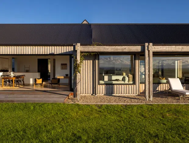 Single-storey contemporary residential home viewed from the lawn, with a broad dark pitched roof, timber-clad facade, extensive floor-to-ceiling glazing, and a deep covered outdoor terrace. The open-plan interior is visible through the glazing, while the terrace includes reclining loungers and built-in timber posts with climbing vegetation. The setting is a landscaped suburban or rural garden in warm late-day light.