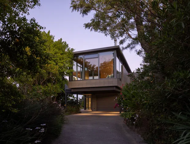Exterior view of a contemporary elevated two-storey home approached by a paved driveway through dense landscaped vegetation. The house has a low-pitched roof, extensive upper-level glazing, a projecting balcony or living area, dark structural framing, and an integrated garage at ground level. The image is captured in soft dusk light.