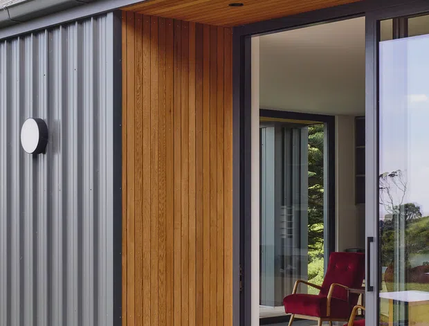 Close-up exterior view of a compact contemporary building with a steeply pitched grey metal roof, vertically ribbed grey metal cladding, warm vertical timber wall panelling, and dark-framed full-height glazed sliding doors. A timber-lined recessed entry soffit opens onto a timber deck, with a red armchair and a neutral-toned interior visible through the glazing. Tall coastal-looking trees surround the structure.