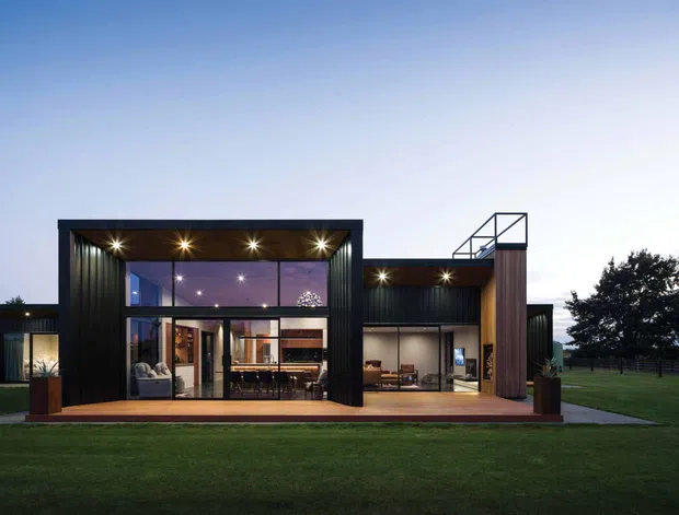 A contemporary single-storey house viewed from the lawn at dusk. The facade combines dark vertical cladding, extensive floor-to-ceiling glazing, warm timber-toned soffits and columns, and a broad timber deck. Interior living and dining areas are visible through the illuminated glass, while a simple rooftop terrace or balcony structure rises above part of the building. The setting includes a large lawn and mature tree.