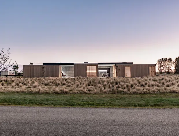 A low, single-storey contemporary residence viewed from across a road at dusk. The elongated building has a flat roof, dark timber-toned cladding, recessed glazed openings, and a central courtyard-like entry zone. It sits within a broad naturalistic landscape of tussock grasses and lawn, with trees framing the property.