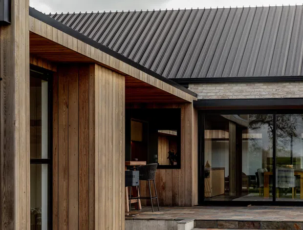 A contemporary low-rise residential building with warm vertical timber cladding, a sharply pitched dark metal roof, exposed timber soffits, and extensive black-framed glazing. The image focuses on a sheltered outdoor threshold and glazed living or dining area, with a glimpse of interior furniture beyond.