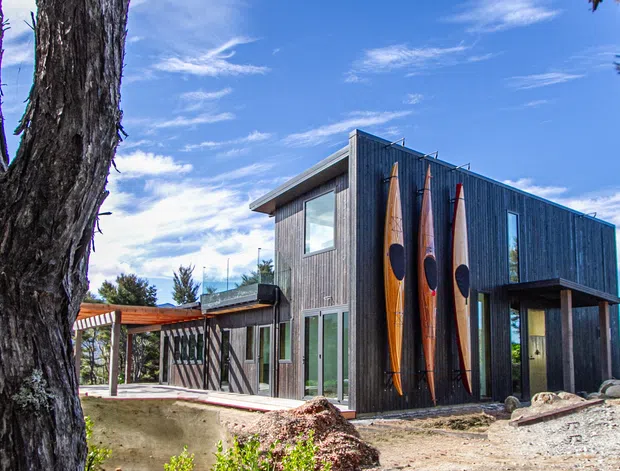 Exterior view of a contemporary two-storey dark-clad house set among mature trees. The home features a simple low-pitched roof, extensive glazing, a covered timber-framed outdoor area, and three vertically displayed surfboards on the facade. Natural rocks and a lightly landscaped ground plane surround the building.
