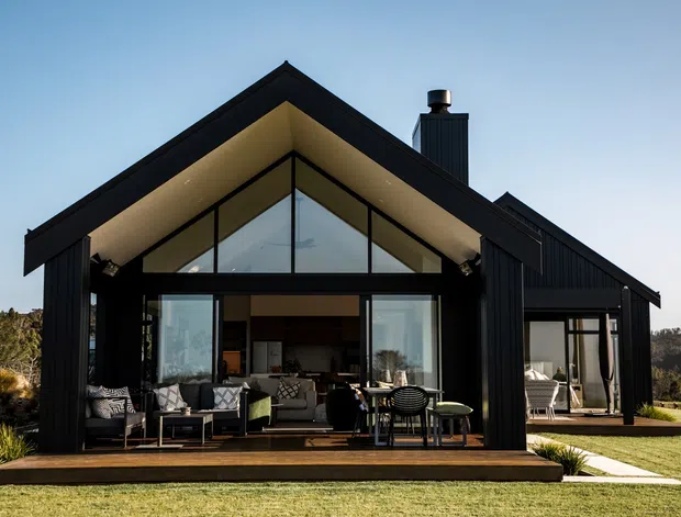 Exterior view of a modern single-storey gable-roof house with bold black vertical cladding, expansive floor-to-ceiling glazing, a covered timber deck, and outdoor seating. The home is set in a landscaped lawn with low planting and a wooded backdrop.
