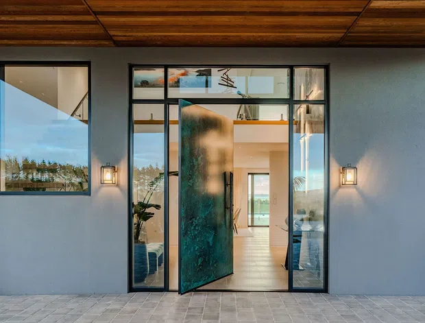 Front entry exterior of a contemporary home, with a broad grey rendered facade, dark-framed glazing, a large pivot entry door, warm wall sconces, and a timber-lined soffit. The open doorway reveals a bright interior beyond, while the foreground is a paved entry terrace.