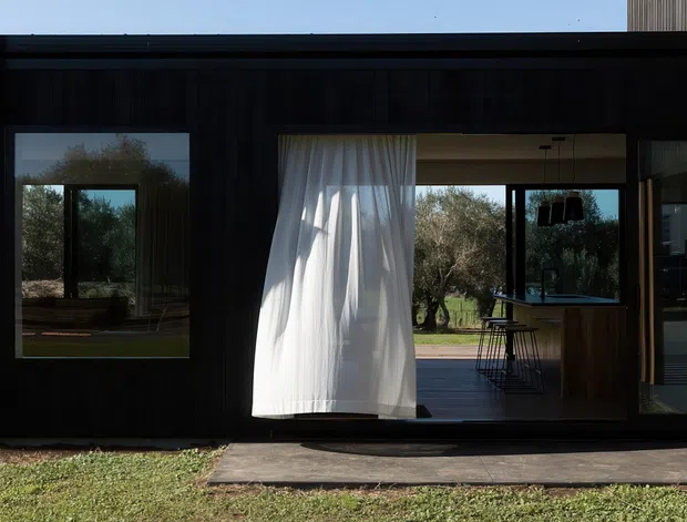 A low, dark-clad contemporary house facade viewed from outside, with expansive black-framed glazing and a large sliding opening. A sheer white curtain hangs in the opening, revealing a timber-toned kitchen or bar area and outdoor deck beyond. The home sits in a landscaped rural or garden setting with mature trees visible through the glazing.