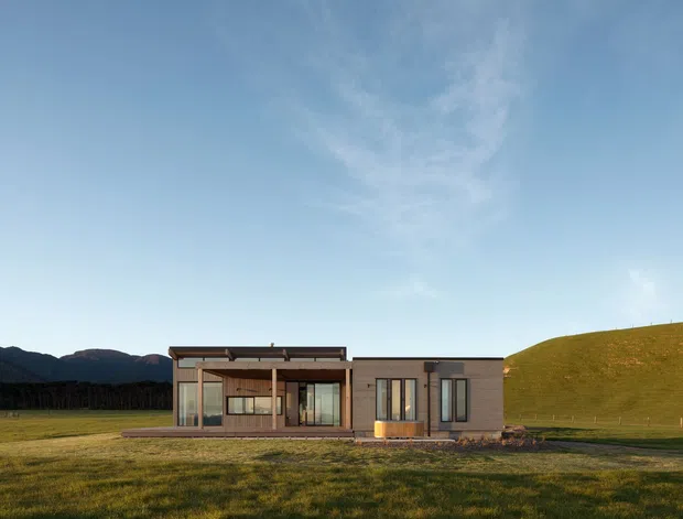A low, single-storey contemporary house set in an expansive rural landscape. The building has a simple rectangular composition with extensive floor-to-ceiling glazing, warm timber detailing, dark metal framing, and pale rendered or concrete-like wall surfaces. A flat or very low-pitched roofline and restrained modern facade contrast with the surrounding grassy field and rolling hills.