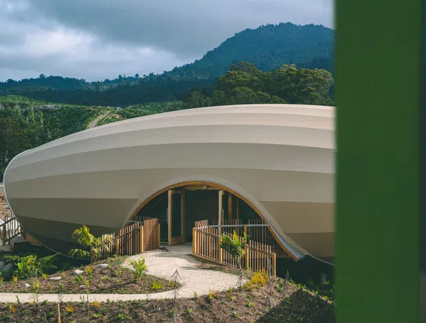 Exterior view of a distinctive sculptural building with a smooth, layered, organic roof and facade forming an arched entrance. Timber-toned railings and entry doors contrast with the pale shell-like structure. The building sits within a landscaped, sloping site with forested hills and mountains in the background, viewed partially through a dark green vertical frame in the foreground.