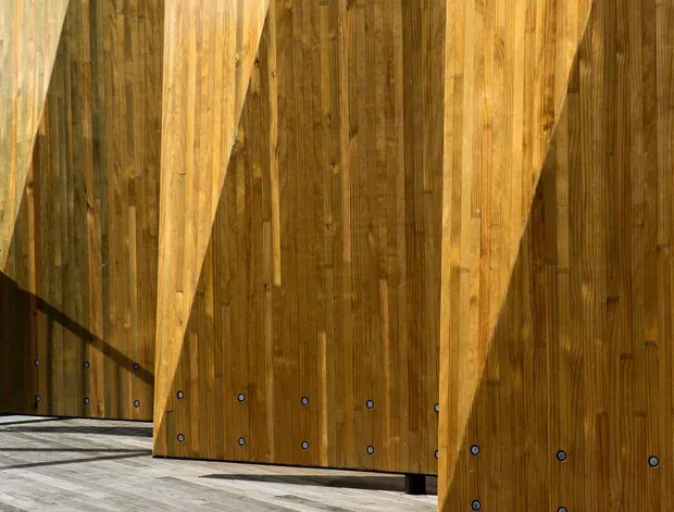 Close-up architectural detail of a series of large angular timber panels or doors mounted above a weathered timber deck. The panels feature vertical wood grain, visible dark fasteners, and dramatic triangular geometric projections or cut-outs, creating strong repetitive shadows and a sculptural facade-like composition.
