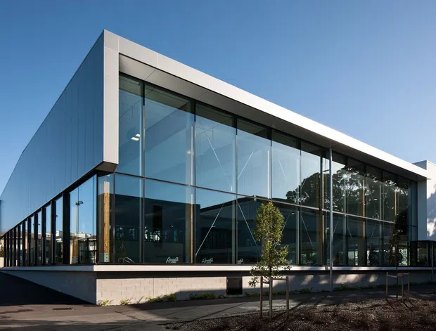 Exterior photograph of a contemporary, single-storey commercial building with a crisp rectangular form, flat roof, extensive floor-to-ceiling glazing, dark slender window frames, and light-coloured solid wall panels. The building sits in a landscaped urban setting with a paved forecourt and young trees.