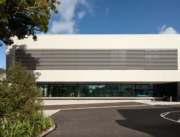 Exterior view of a contemporary institutional or commercial building with a long, low rectangular form. The upper facade is clad in pale horizontal ribbed panels and incorporates a wide band of dark horizontal louvres, while the ground floor has extensive dark glazing. A black-clad entrance volume sits at the right. The building fronts a paved asphalt forecourt with marked parking or access areas, with established landscaping and trees at the edges.