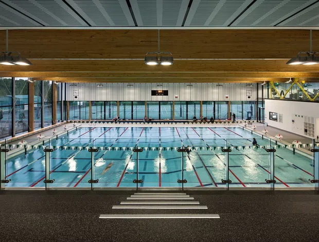 Wide interior view of a large modern indoor aquatic centre, looking across multiple swimming lanes from a raised spectator or circulation area. The pool hall has expansive glazing, timber-clad structural beams, a patterned suspended ceiling, bright overhead lighting, tiled pool surrounds, lane ropes, starting blocks and spectator seating.