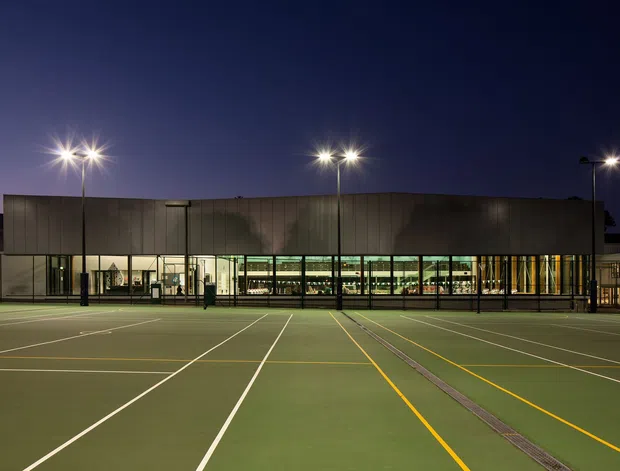 Night-time exterior view of a contemporary institutional or commercial building behind a large illuminated multi-court sports surface. The building has a long, low rectangular form with dark vertical cladding, extensive ground-level glazing, and bright interior lighting. Tall floodlights illuminate the marked green courts in the foreground.