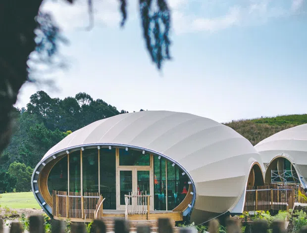 Exterior view of distinctive rounded pod-like buildings with white arched membrane roofs, extensive curved glazing, timber-framed entrances and small timber decks. The structures sit in a landscaped rural or semi-rural setting, viewed through a foreground fence and foliage.