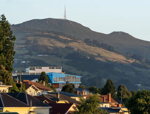 Exterior view across a suburban residential neighbourhood toward a dramatic steep, forested and partly exposed mountain ridge. Foreground homes have pitched roofs, varied roof cladding, brick chimneys and colourful facades, while a contemporary multi-level building with extensive glazing sits among the houses. A communications mast is visible on the mountain summit.
