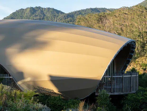 Exterior view of a distinctive curved, tensile-looking architectural roof or shell enveloping a building, set against densely forested mountainous terrain. The structure has a smooth warm beige exterior surface, dark structural edges, vertical metal screening, and arched openings partially visible at ground level.