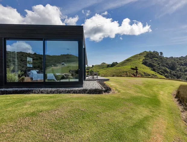 A modern, single-storey dark-clad structure with extensive floor-to-ceiling glazing sits beside a carefully maintained lawn on a sloping, hilly rural or bush setting. The glass reflects and reveals a simply furnished interior, while a gravel perimeter and small paved outdoor area connect the building to the landscape. Bright blue skies and rolling green hills create a spacious natural context.