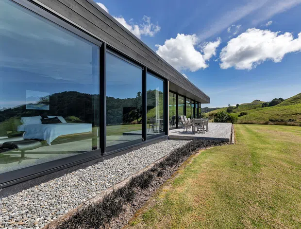 Single-storey contemporary residence set in a rolling rural landscape, viewed from outside along a long glazed facade. The building features extensive floor-to-ceiling sliding glass doors in dark frames, horizontal dark weatherboard cladding, a low-pitched roofline, and a gravel strip beside the glazing. A dining terrace extends from the building onto a broad lawn, while a bedroom and furnishings are visible through the glass.