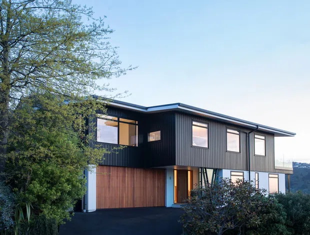 Exterior view of a contemporary two-storey house with dark vertical cladding, a low-pitched roof, expansive rectangular windows, and a warm timber garage door. The home sits on a landscaped suburban site with mature trees and shrubs, and appears to overlook a distant hilly landscape.