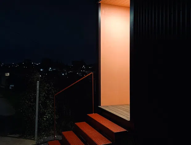 A dramatic nighttime exterior view of a contemporary dark-clad building, featuring a warmly illuminated recessed entry or porch with a timber-lined soffit and deck. A series of red-orange tiled steps leads up from a concrete walkway, bordered by dark landscaping and a simple metal handrail. Distant residential lights are visible beyond the site.