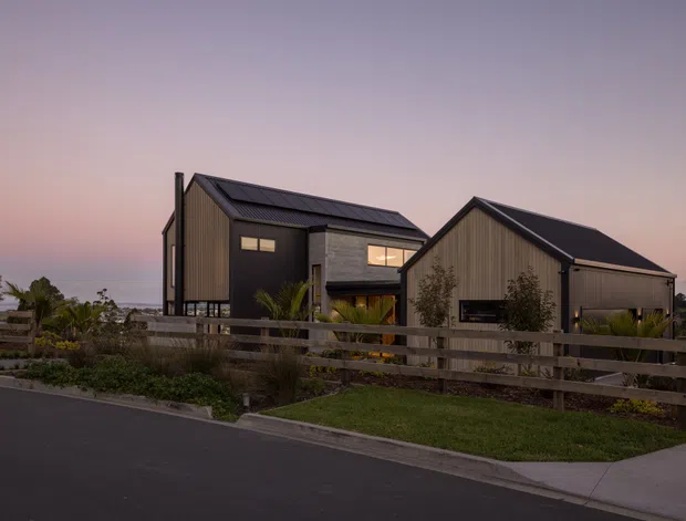 Exterior view of a contemporary multi-building residence at dusk, with dark vertical cladding, pitched metal roofs, a central concrete-toned wall, and warm illuminated windows. The home sits behind a horizontal timber fence with landscaped planting and a paved driveway or walkway in the foreground.
