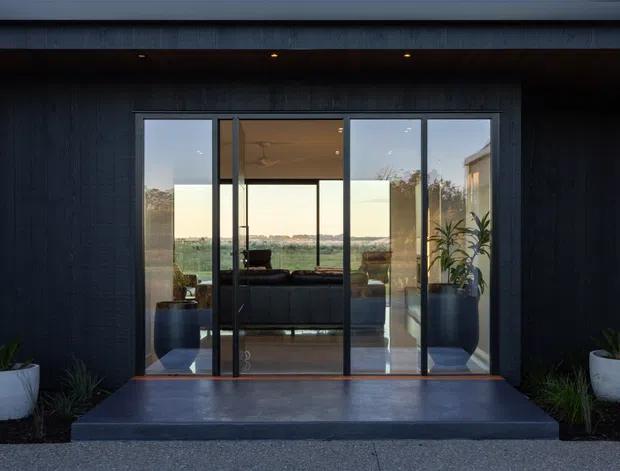 Front exterior view of a contemporary dark-clad building entry, featuring a broad aluminium-framed glazed door and sidelights opening into a living room. A covered soffit with recessed downlights, raised dark entry steps, and symmetrical potted planting frame the entrance. The glazing reveals a sofa, ceiling fan, and expansive landscape beyond.