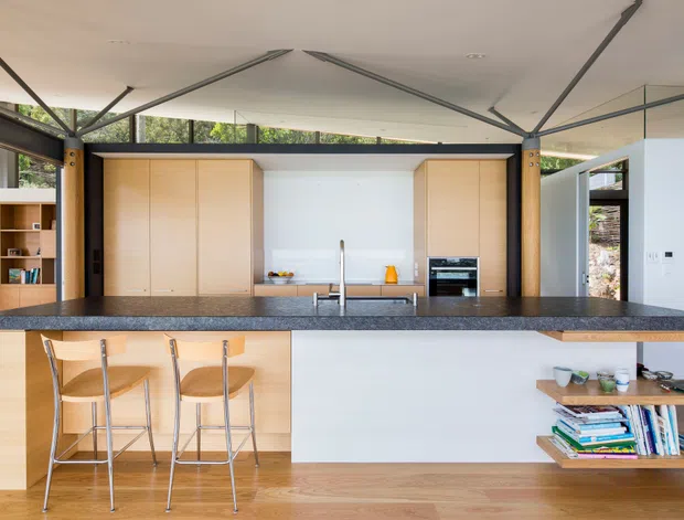 A bright, open-plan contemporary kitchen with a large dark stone island and integrated breakfast bar seating. Light timber cabinetry frames the rear cooking zone, with a stainless-steel sink and tap, built-in oven, and open shelving. Exposed dark structural posts and beams, extensive glazing, and a white geometric ceiling create a spacious, modern interior. A low timber display shelf with books and decor sits at the right.