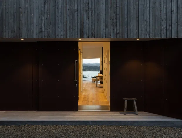 A dark, minimalist building exterior with a heavily textured vertical timber upper facade and near-black lower wall panels. A central open doorway reveals a warm timber-floored interior with a small table and chair, framed by a scenic view toward water and hills. A simple timber stool sits on the covered threshold, with gravel in the foreground.