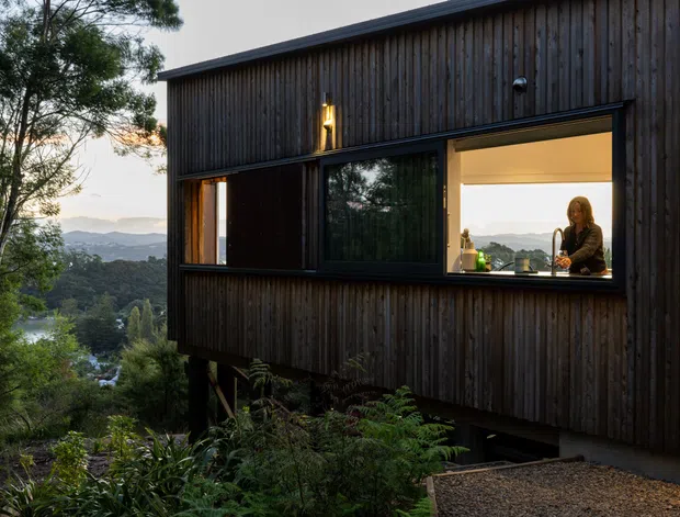 Exterior dusk view of a contemporary elevated house clad in dark vertical timber boards, with large glazed openings glowing warmly from within. The building sits among dense vegetation on a sloping site, with layered hills visible beyond. A person is visible inside at a kitchen or living area counter.
