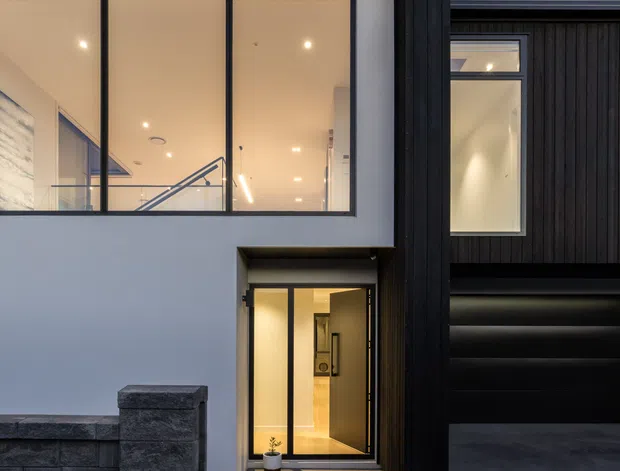 Contemporary two-storey residential facade at dusk, featuring a crisp white rendered volume paired with dark vertical timber cladding. Large black-framed windows reveal a warmly illuminated stair and entry interior, while a recessed glazed front door opens onto a paved path bordered by white gravel. A dark garage and low masonry boundary wall complete the facade composition.